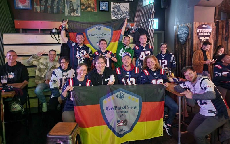 Eine Gruppe von GPC-Mitgliedern in einem Gruppenbild. Die Fanclub-Fahne ist einmal vorne und einmal hinten gespannt. Sie sitzen in einem Pub in London.