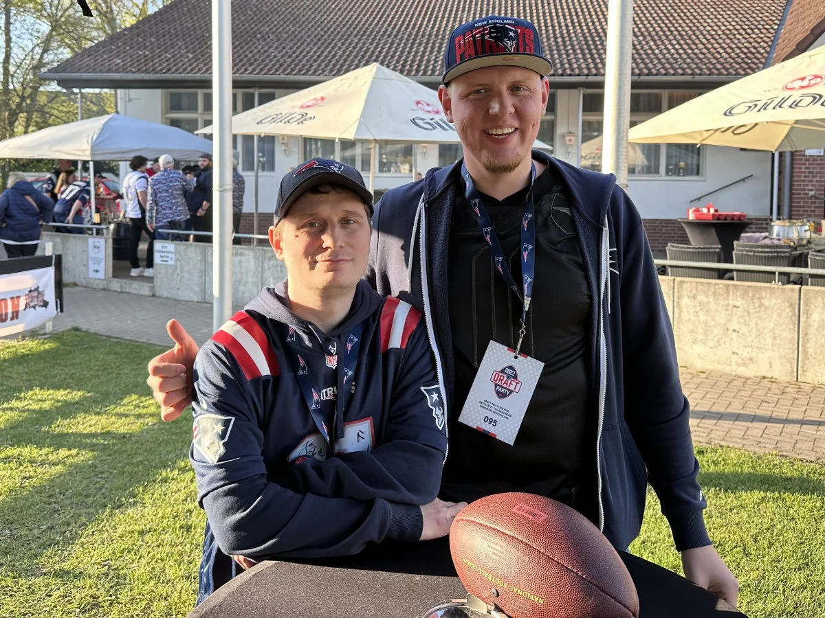 Zwei Mitglieder im Patriots-Gear stehen nebeneinander, vor ihnen liegt ein Football.