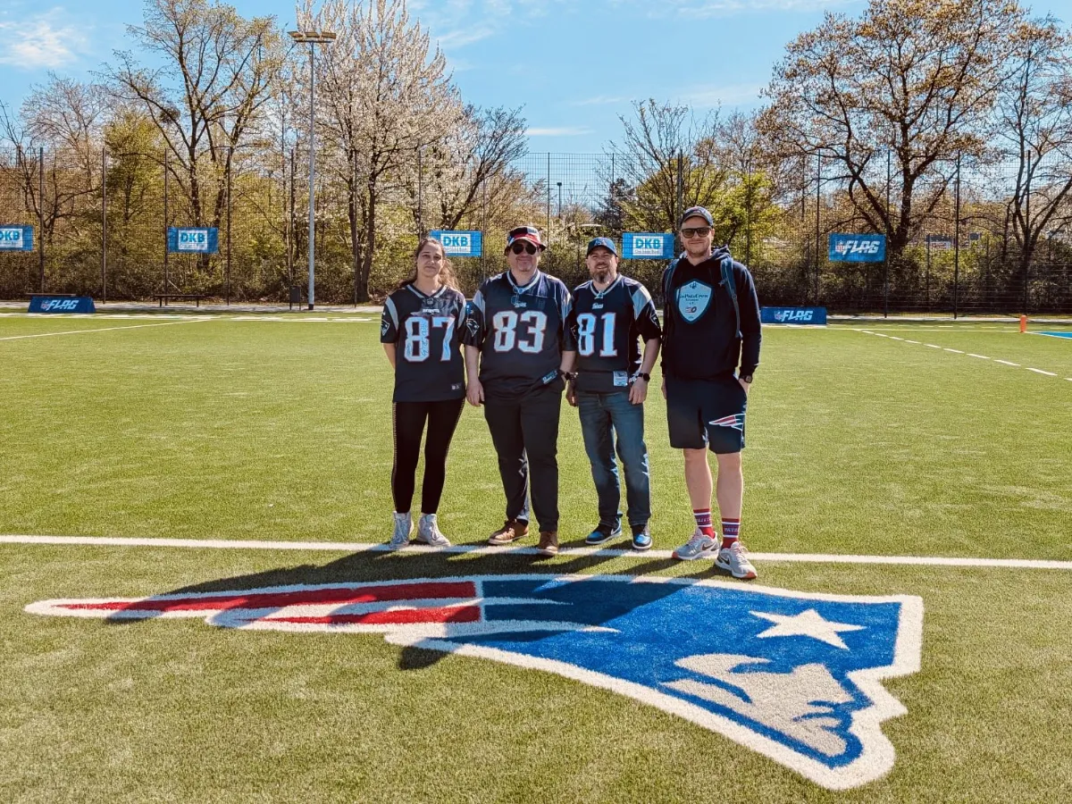Vier Mitglieder im Patriots-Gear stehen auf einem Football-Feld, auf dem Boden ein großes Patriots Logo