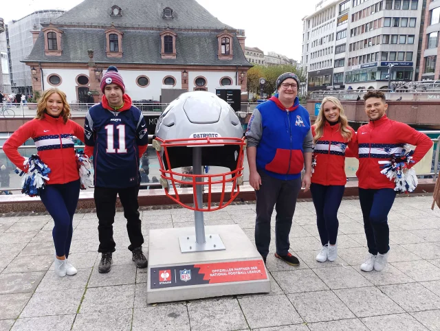 Zwei Mitglieder stehen gemeinsam mit mehreren Cheerleadern neben einem überdimensionalen NFL-Helm.