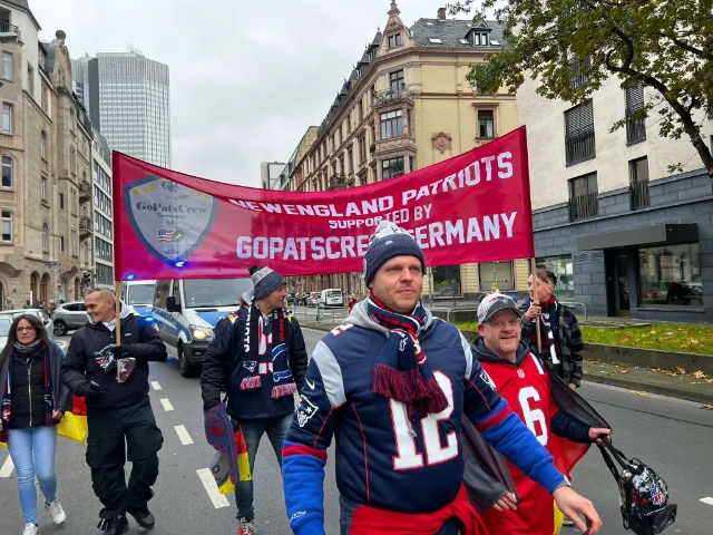 Sechs Mitglieder beim Fanmarsch in Frankfurt am Main. Sie halten ein rotes Banner mit der Aufschrift "New England Patriots - supported by GoPatsCrew Germany und dem Fanclub Logo