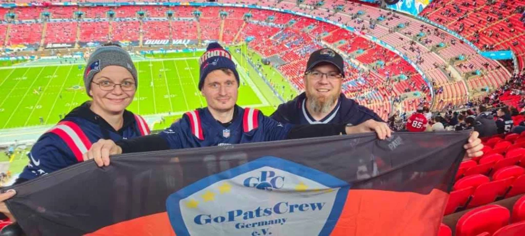 Drei Mitglieder stehen im Wembley Stadion, im Hintergrund sind die Trib&uuml;nen zu sehen