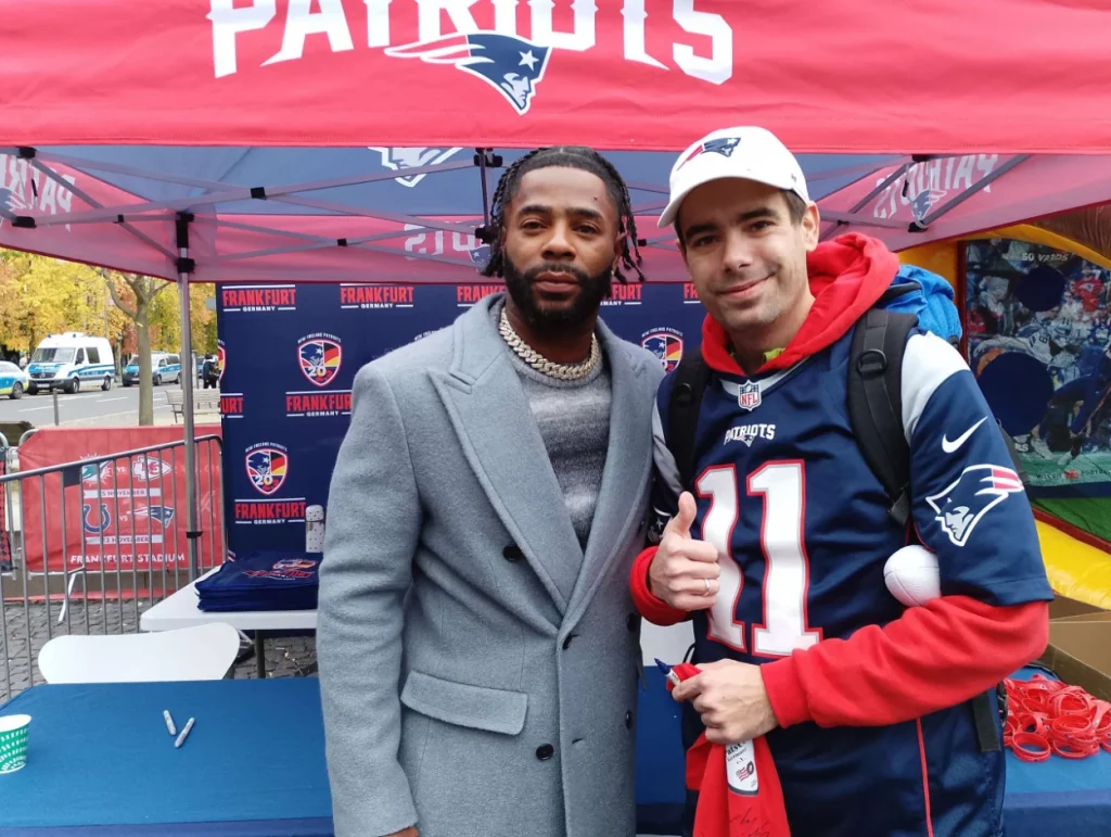Ein Mitglied steht neben einem Football-Spieler, im Hintergrund ein Patriots-Stand